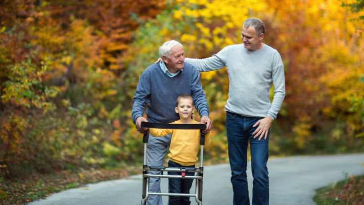 A grandfather in a walker walking with support from his son and grandson on a trail surrounded by fall leaved trees.