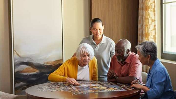 Residents at The Landing at Watermere Frisco work on a jigsaw puzzle at a round table with a caregiver standing nearby, offering support in a warm, sunlit room.