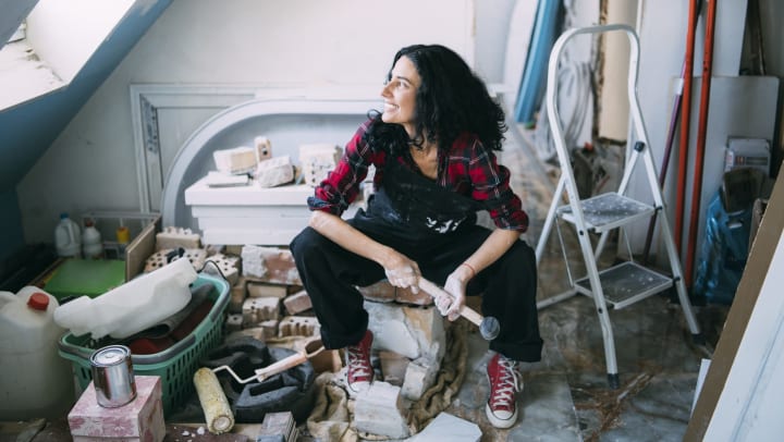 Smiling woman with mallet in hand surrounded by painting supplies and loose bricks. 