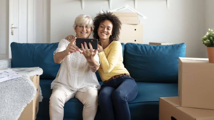 Two smiling women taking a selfie on a camera phone while sitting on a blue couch in an emptied house surrounded by moving boxes
