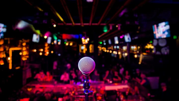 Point of view of a person on a stage. A mic is in the foreground. A dark, expansive nightclub is in the background.