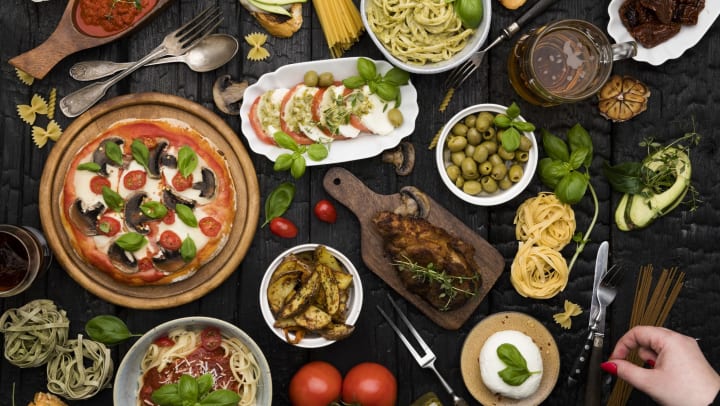 View from above of a variety of Italian food dishes including a pasta bowl, bruschetta, and a margherita pizza, all set on a black table. 