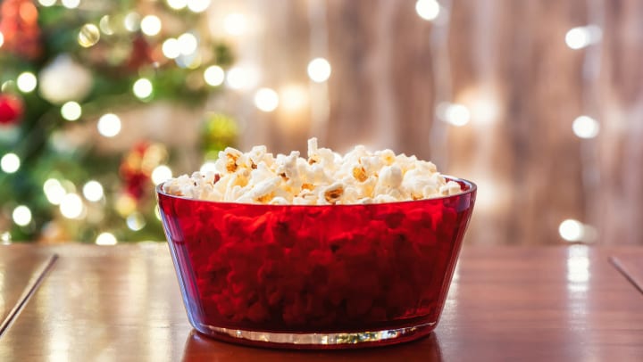 A red bowl filled with popcorn sitting on a table with a Christmas tree and festive lights in the background