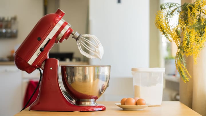 Red table mixer on wood counter. Plate of eggs and container of flour with yellow flowers hanging above them.