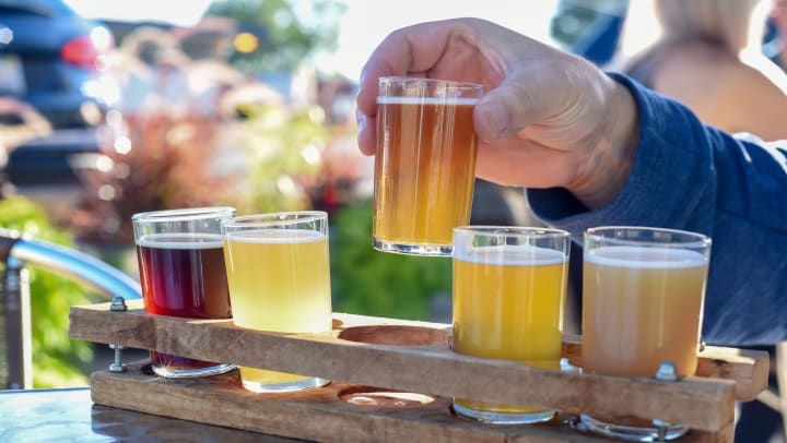 View of a beer flight sitting on an outdoor table, with a hand lifting one of the glasses.