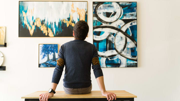 Back view of a young man sitting on a bench and admiring the various paintings on the wall of an art gallery.