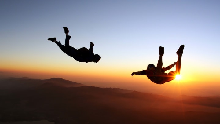 Two people skydiving with the sun peeking out over the mountains in the background
