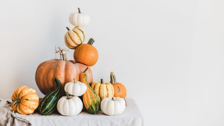 An assortment of white, orange, and green pumpkins stacked on a table with a soft grey tablecloth against a neutral grey background