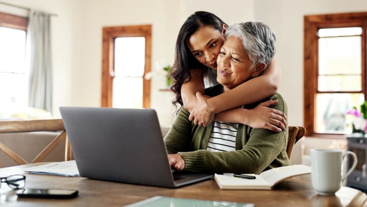 Daughter hugging mother, both smiling while looking at a laptop