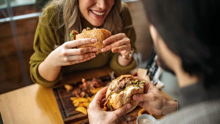An over-shoulder view of a man and woman sitting across from each other at a table, both holding hamburgers on sesame buns | burgers in Allen
