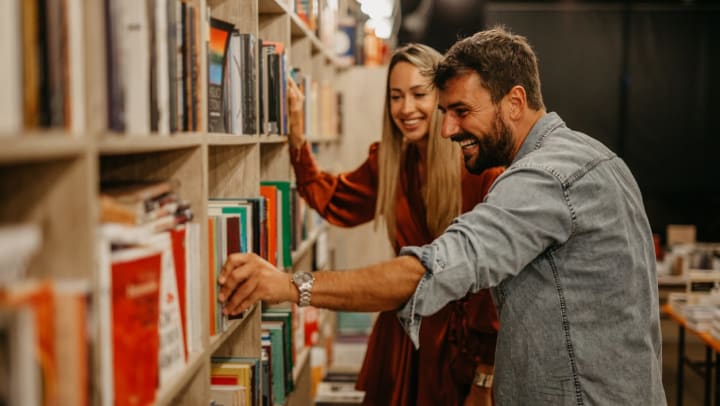 A man in a blue denim shirt and a woman in an orange dress both smile as the man reaches toward a long row of bookcases to pull a title from the shelf at one of the local bookstores in Tampa