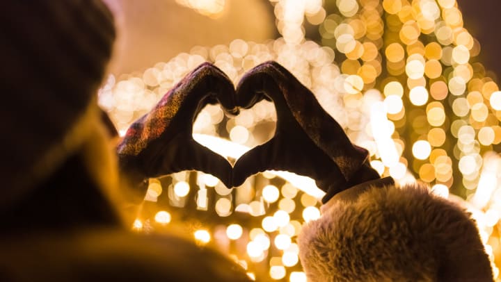 Woman making a heart shape with her hands in mittens towards festive lights