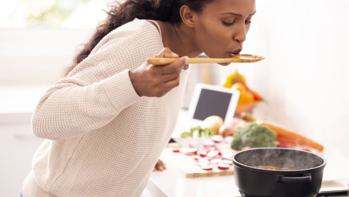 A woman tastes the soup that is simmering on her stove. Chopped vegetables sit on the counter in the background.