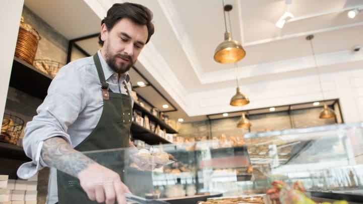 A man wearing an apron using tongs to grab a baked good from a display case.