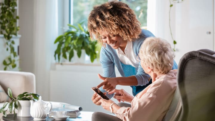 A young woman smiling and pointing at a smartphone while standing next to a seated older woman