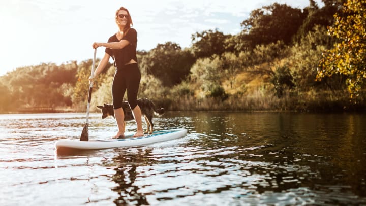 A smiling woman enjoys a peaceful moment on the water with her dog on a paddle board.