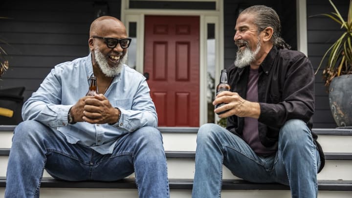 Two senior men holding bottles,  smiling, and talking on a front porch