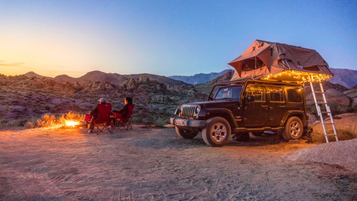 Two people sitting on camping chairs next to a jeep with a pop-up tent atop it.