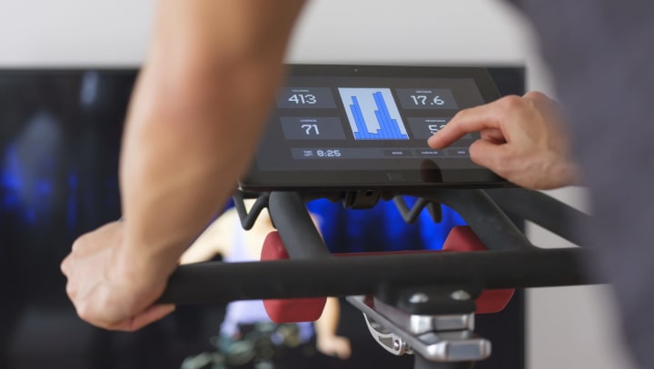Close-cropped view of a person on a stationary bike with a digital display and a television monitor with a fitness trainer in the background