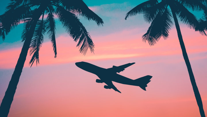 Two palm trees outlined against sunset with an airplane flying in between in the background.