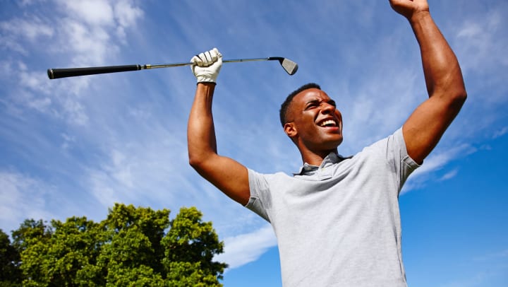 Smiling young man holding a golf club with his arms raised over his head with blue sky and green trees in the background