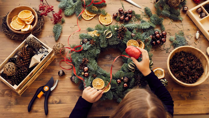 Overhead view of a woman making holiday decorations using greenery, pine cones, ribbon, and dried orange slices. 