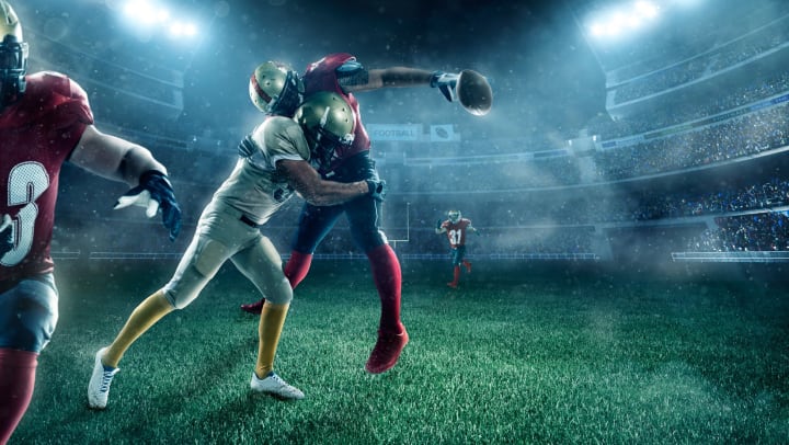 A quarterback being hit by another player, the ball falling out of his hand. A well-lit stadium in the background. It’s a night game. 