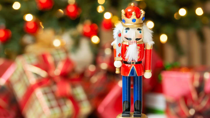 A nutcracker with a blurred lighted tree and wrapped gifts in the background.