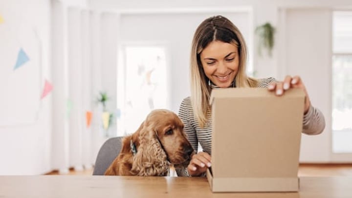 A smiling woman opening a brown box with her dog sitting next to her.