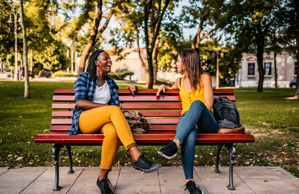 Two friends chatting on a park bench under lush trees at The Grove in Battle Creek, Michigan