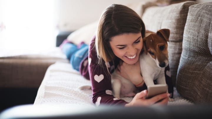 Woman lies on the couch with a dog in her arms while both look at a phone in her hand.