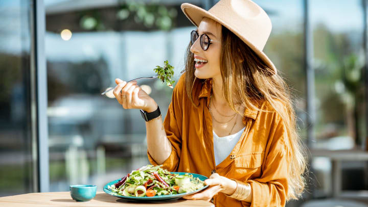 A woman in a hat holding up a fork with lettuce on it and smiling while holding a plate with a salad on it. 