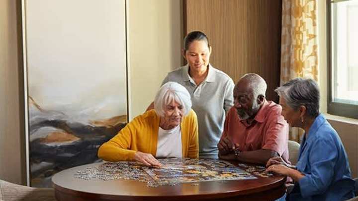 Residents at The Landing at Watermere Woodland Lakes work on a jigsaw puzzle at a round table with a caregiver standing nearby, offering support in a warm, sunlit room.