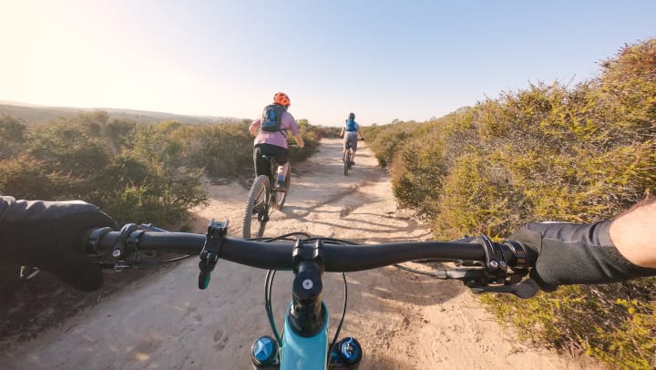 A first-person view from a mountain bike, looking ahead from behind handlebars at a trail and two mountain bikers ahead