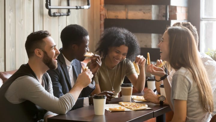 A group of women and men sit around a table eating pizza and drinking coffee.