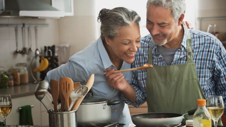 Older man and woman smiling and cooking over a skillet in a kitchen