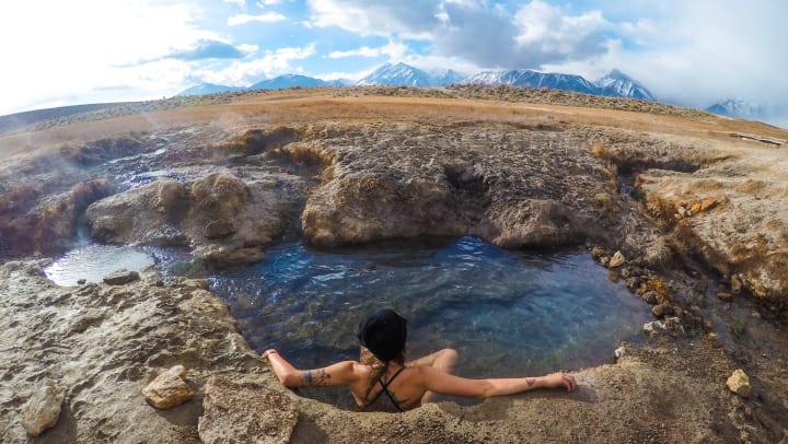 A woman in a bathing suit sitting in a natural hot spring with snow-covered mountains in the distance