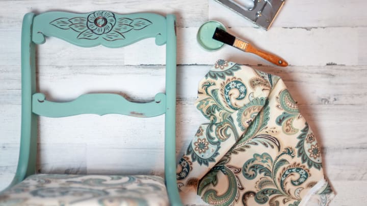 A freshly painted and reupholstered dining chair sits next to a paint brush, paint, and a fabric swatch. 