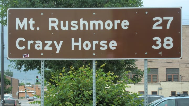 A street sign with the words, “Mt. Rushmore 27 Crazy Horse 38”.