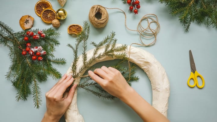Hands holding a pine branch to a wrapped wreath with various craft supplies spread out around them. 