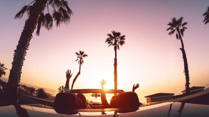 Image of two people in a convertible car driving with palm trees in the background