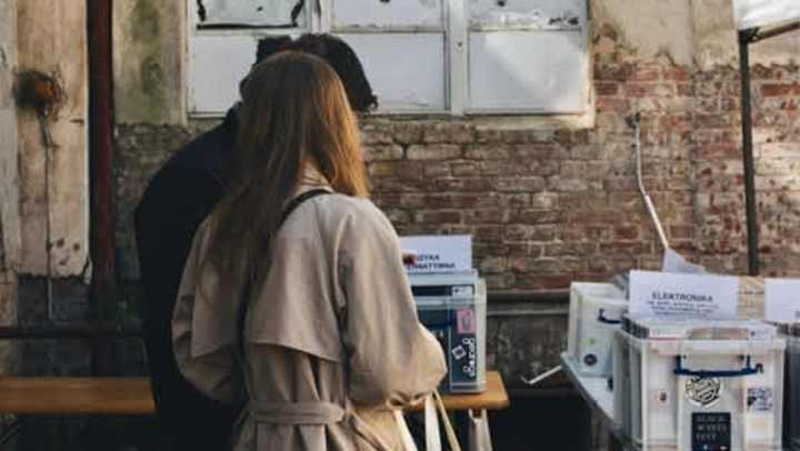 A man and a woman stand with their backs to the camera, looking down at items on a table at an outdoor market or fair. The woman wears a tan trench coat. They are standing against a background of an old brick wall with a dilapidated, white-framed window above. The table in the foreground holds several white plastic bins.