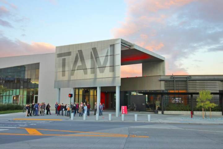 An outside view of the Tacoma Art Museum from across the street. A group of people stand outside of the museum, with a large concrete exterior and the letters TAM on the building. 