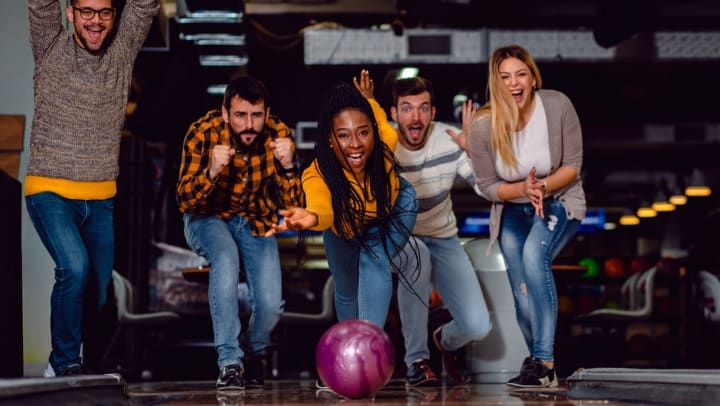 a woman rolling a bowling ball onto the lane while friends cheer behind her | Bowling alleys near Irvine