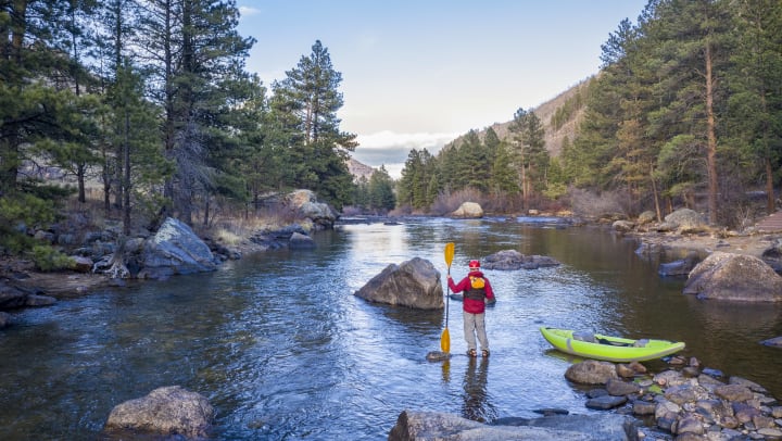 Man standing on a rock in a river, holding a paddle in his left hand with a kayak to his right.