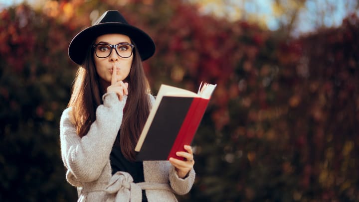 Woman in glasses and a hat putting a finger to her lips while holding a book.