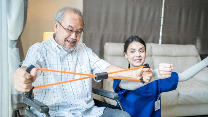 A woman in blue scrubs with her arms up looking at an elderly man in a wheelchair stretches a resistance band.