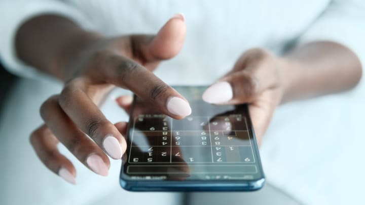 A person is wearing a white outfit, using both of their hands, painted with light pink nail polish is holding an iPhone, playing a game of Soduku 
