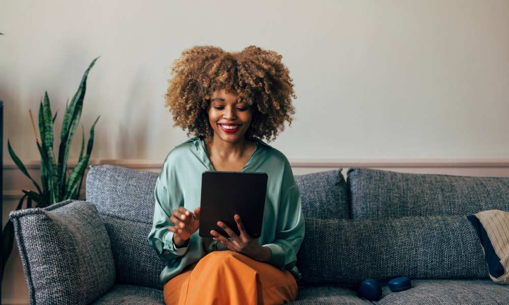 Woman smiling and using a tablet while sitting on a cozy couch with plants nearby at Crestwood Manor in Frederick, Maryland