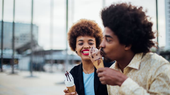 A young man taking a bite of an ice cream cone as a young woman holding a cone watches and smiles. 